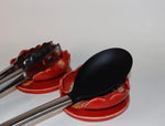 Black spoon and tongs on red ceramic spoon rest with a white background