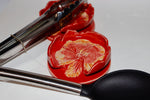Red ceramic spoon rest with floral design and metal tongs on a white background