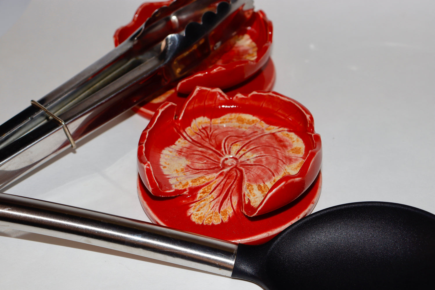 Red ceramic spoon rest with floral design and metal tongs on a white background