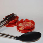 Red ceramic spoon rest with floral design and tongs, alongside a black spoon on a white background